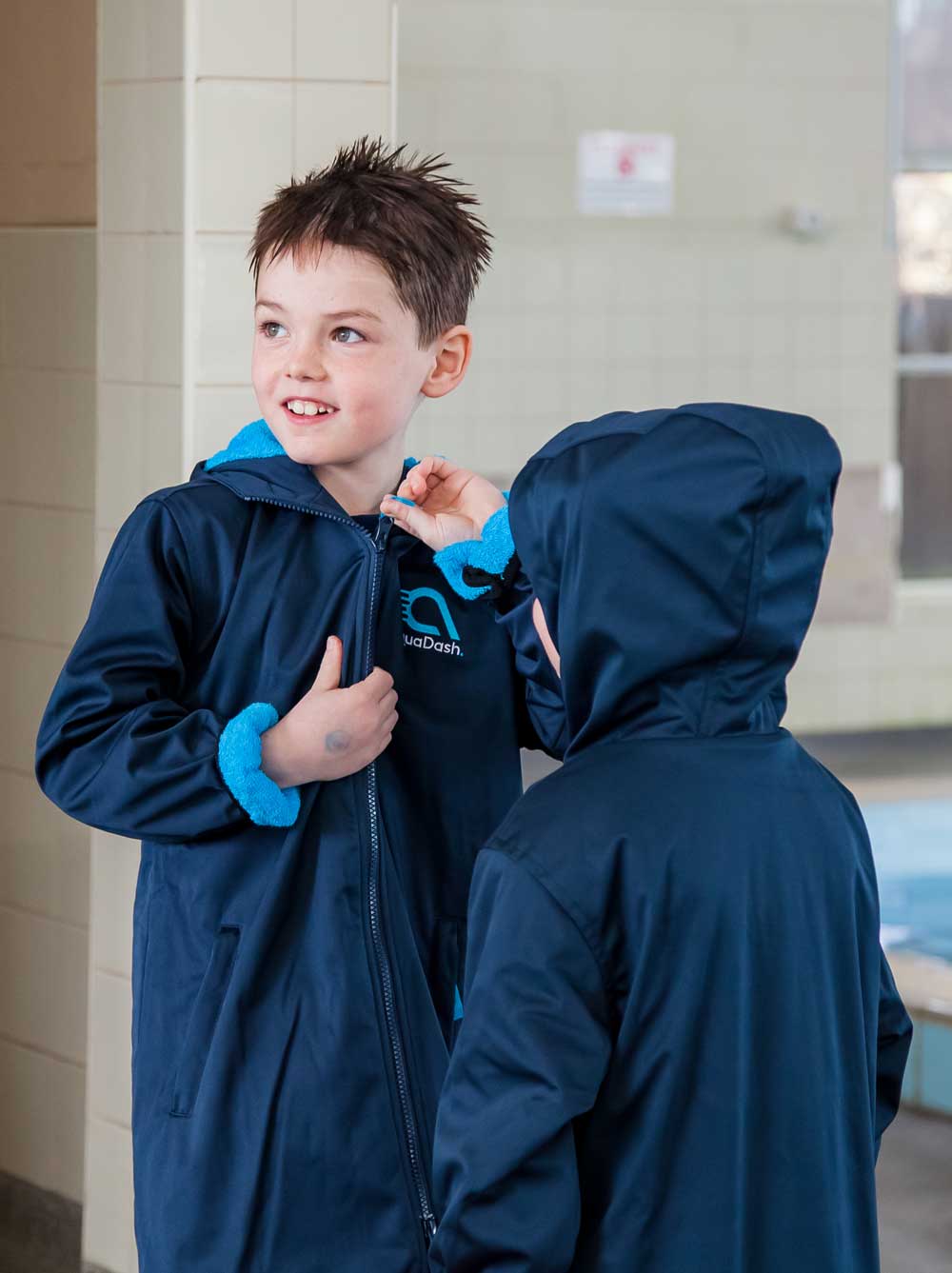 A boy zipping up a swim parka next to a swimming pool and standing with a sibling. They are both warm and dry in their towel lined swim coats